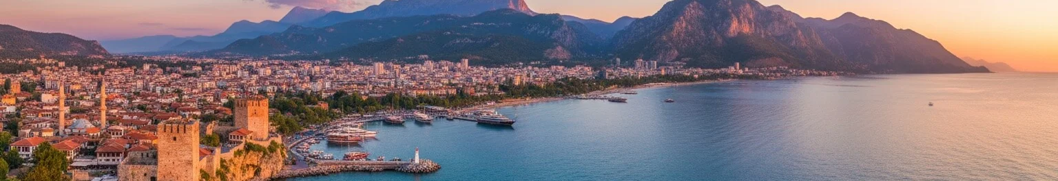 Antalya'nın tarihi Kaleiçi limanı ve şehir surlarının çok geniş panoramik görünümü, turkuaz Akdeniz'de yanaşmış tekneler ile, görkemli dağların arkasında renkli gün batımı gökyüzü.
